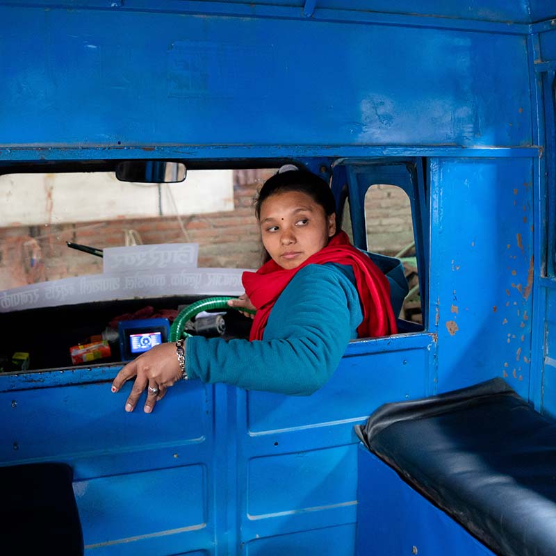A woman sits in the driver's seat of a safa tempo truck, and looks back through bright blue interior of the cab.