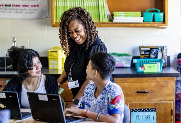 A teacher smiles at two students sitting front of devices.