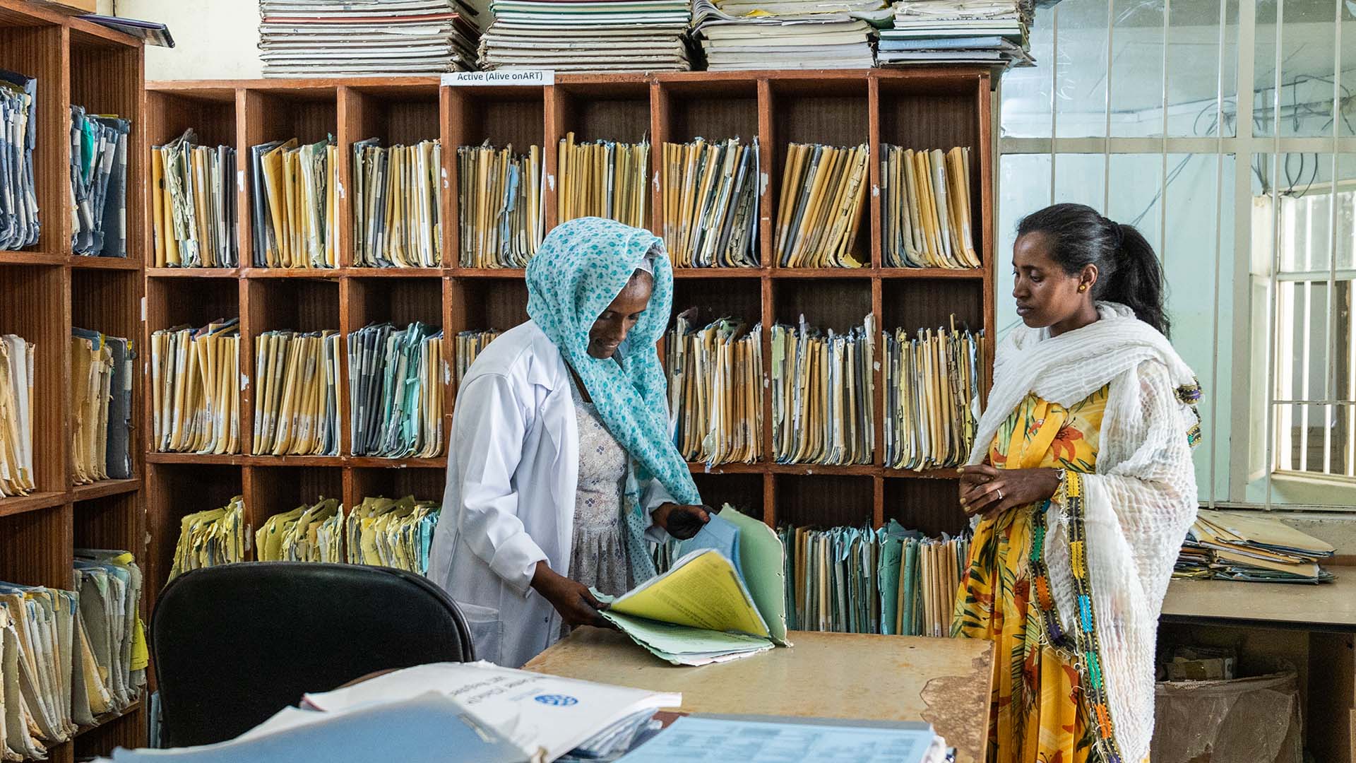 Two women stand in front of a wall of files. One woman looks through some papers.
