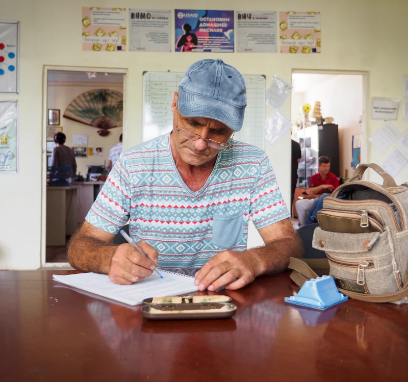 A man sits at a table and writes on a clipboard.