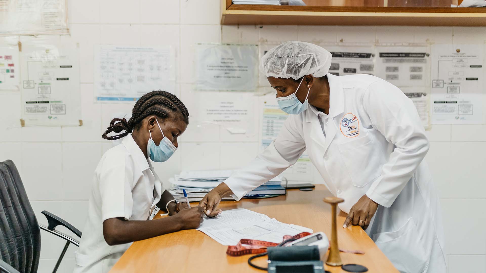 Two nurses wearing face masks and white coats work on a written report.