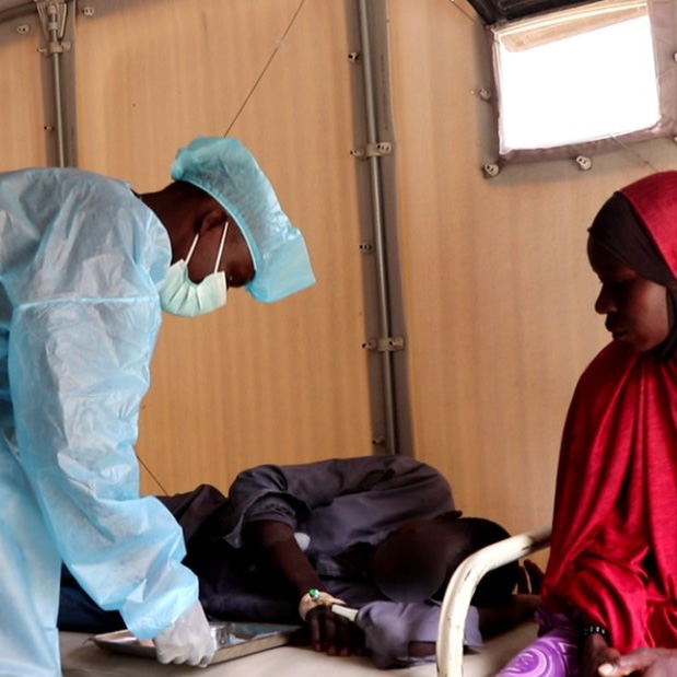 A medical worker in personal protective equipment tends to a patient laying on a bed.