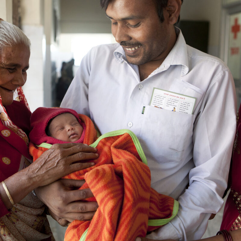 A mother and grandmother looks at a baby being held by a father.