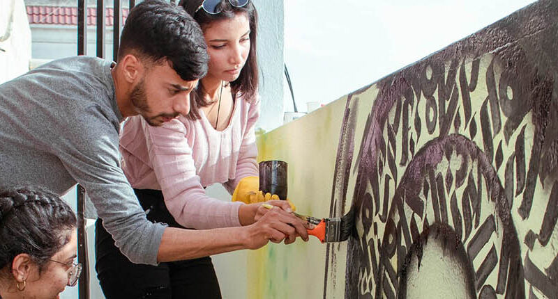 Three young people paint a mural on a low concrete wall.