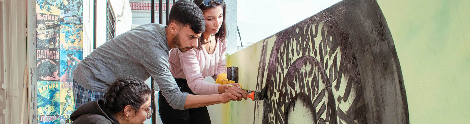 Three young people paint a mural on a low concrete wall.