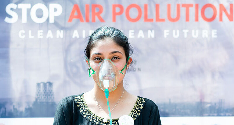 A woman wearing an oxygen masks poses in front of a sign that says "stop air pollution."