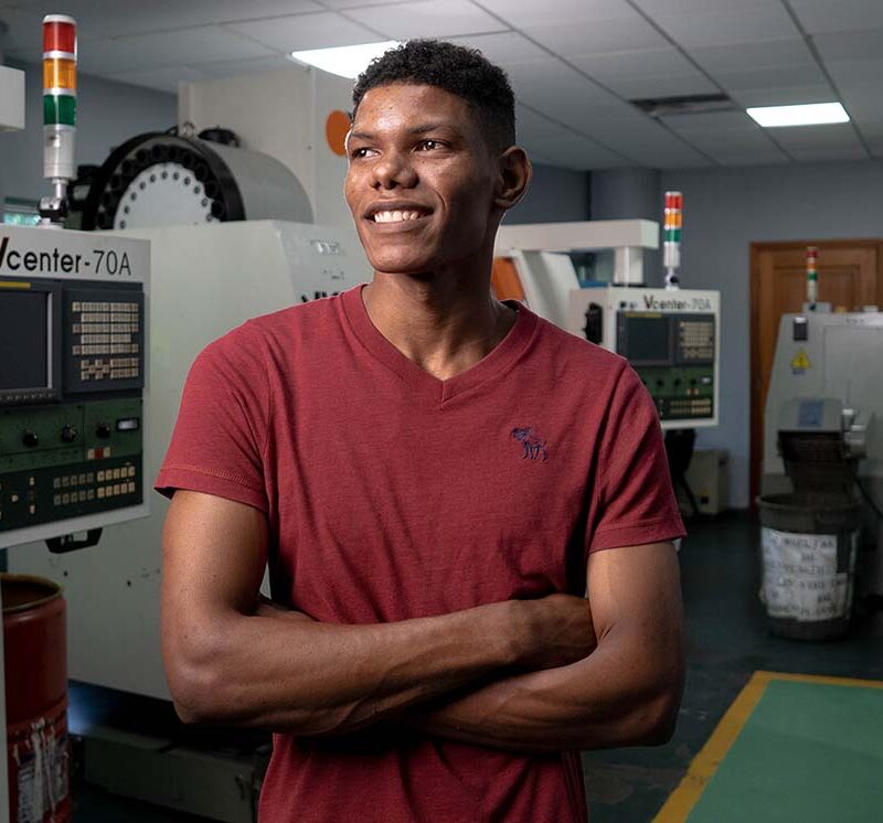 A young man in a red t-shirt stands with his arms folded in a manufacturing lab.