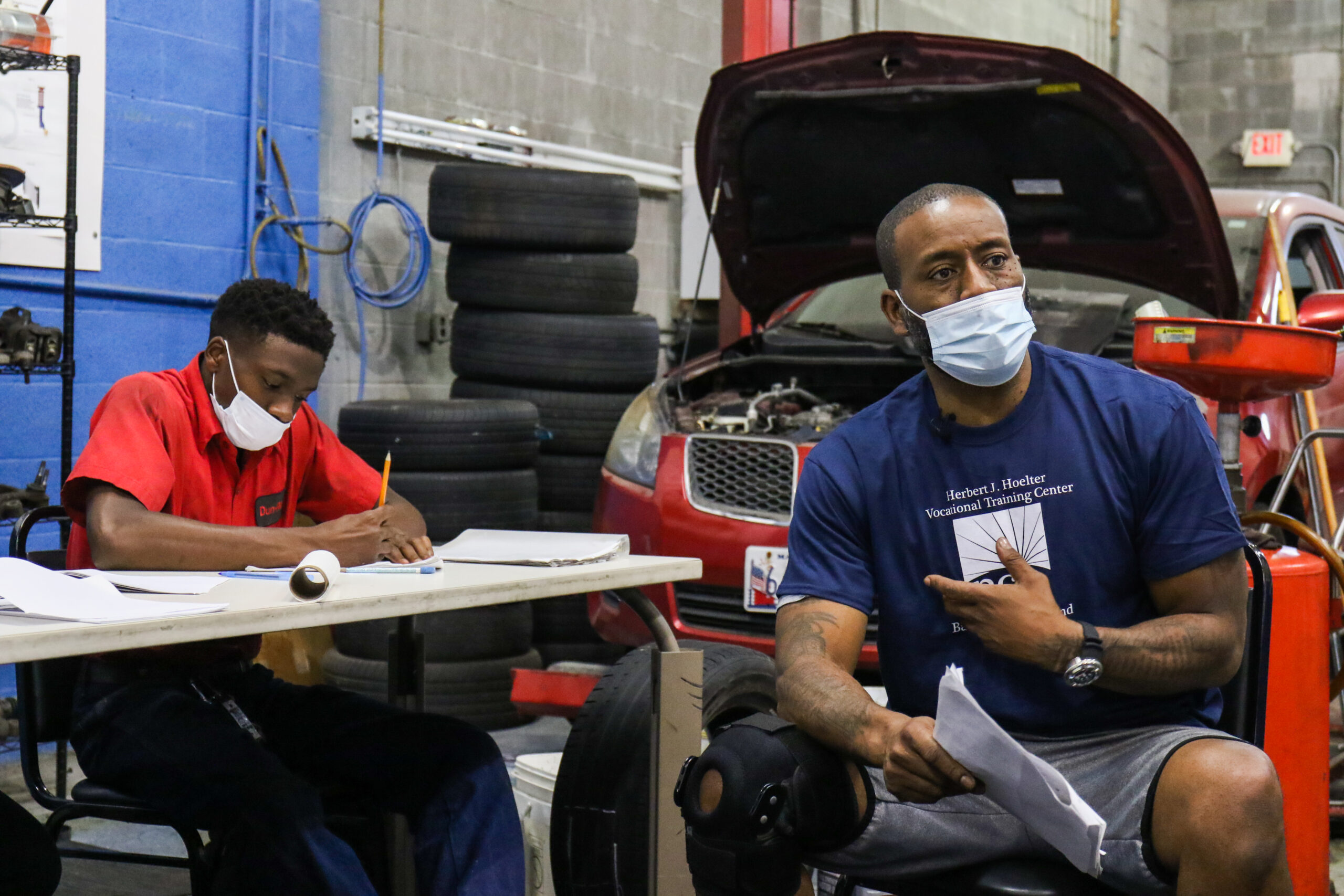A man in a blue NCIA-branded t-shirt sits surrounded by students and vehicles in his mechanic's classroom.