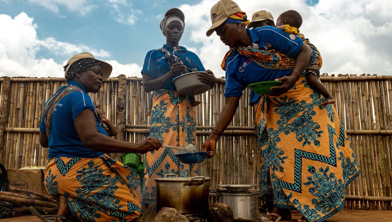A group of women wearing babies and FHI 360 polo shirts serve nutritional porridge that's being cooked over an open fire.