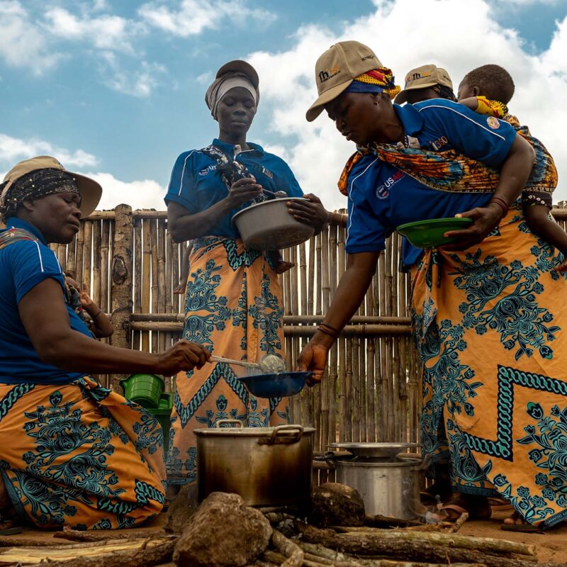 A group of women wearing babies and FHI 360 polo shirts serve nutritional porridge that's being cooked over an open fire.