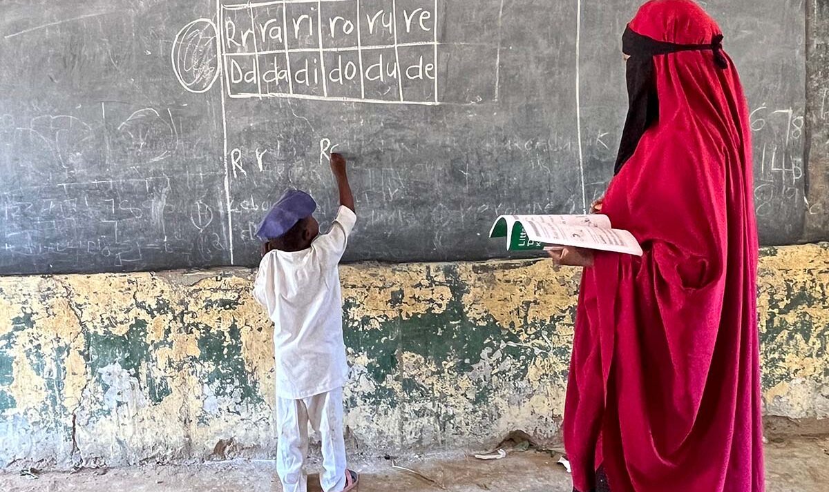 A young boy writes on a chalkboard while his teacher stands behind him and monitors his work