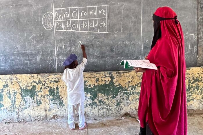 A young boy writes on a chalkboard while his teacher stands behind him and monitors his work