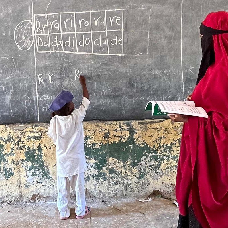 A young boy writes on a chalkboard while his teacher stands behind him and monitors his work