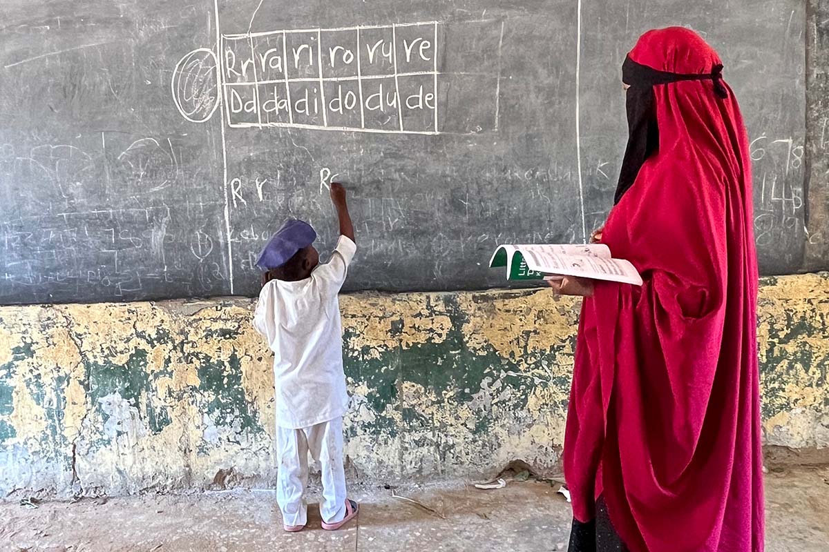 A young boy writes on a chalkboard while his teacher stands behind him and monitors his work