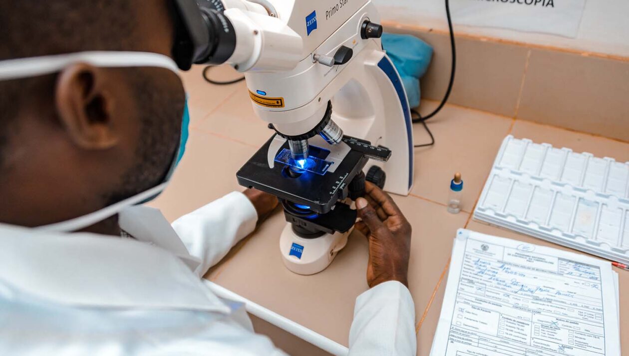 A man wearing a medical mask and lab coat looks into a microscope