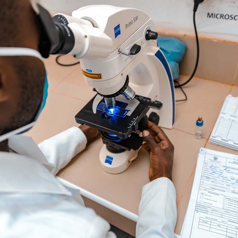 A man wearing a medical mask and lab coat looks into a microscope