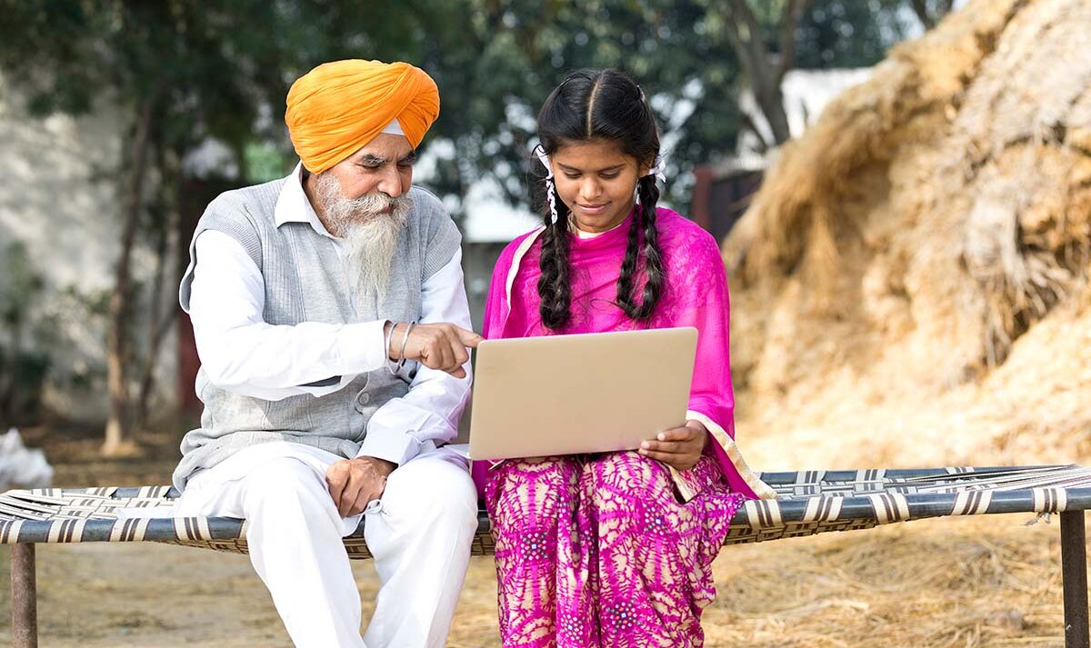 A grandfather wearing white clothes and a bright orange turban sits next to a young girl wearing a bright pink sari while they both look at a laptop