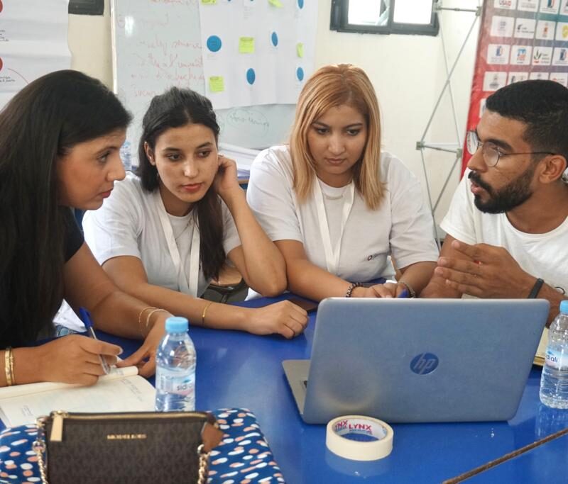 3 young people sit at a blue table with a woman discussing something while looking at a laptop computer