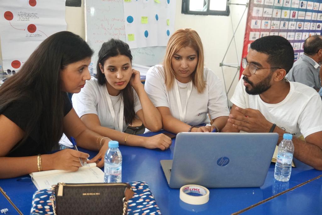 3 young people sit at a blue table with a woman discussing something while looking at a laptop computer