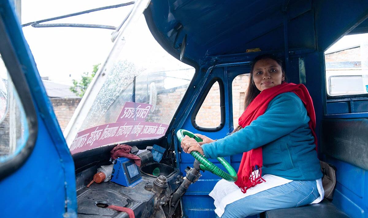 A woman wearing a blue top and red scarf sits behind the wheel of a three-wheeled vehicle