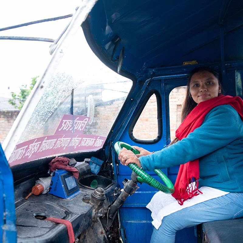A woman wearing a blue top and red scarf sits behind the wheel of a three-wheeled vehicle