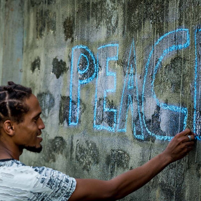 A man smiles while writing PEACE in block letters on a wall with blue chalk