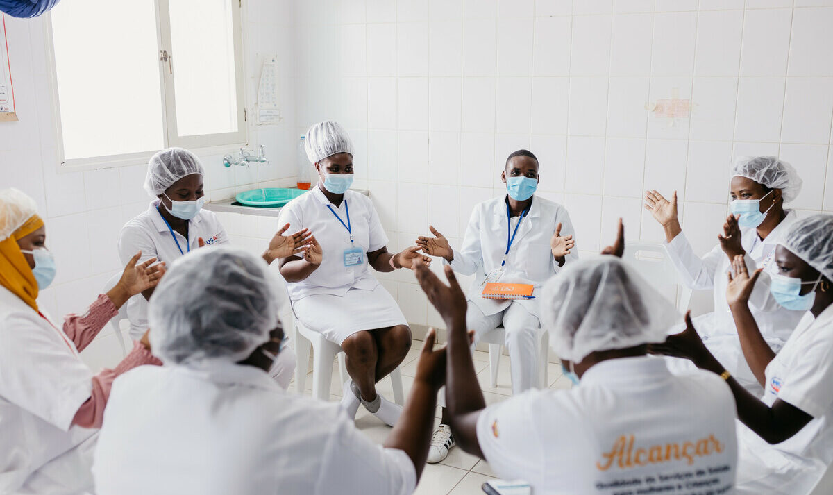 A group of nurses wearing face masks and white dresses with "Alcancar" printed on the back sit in a circle and clap.
