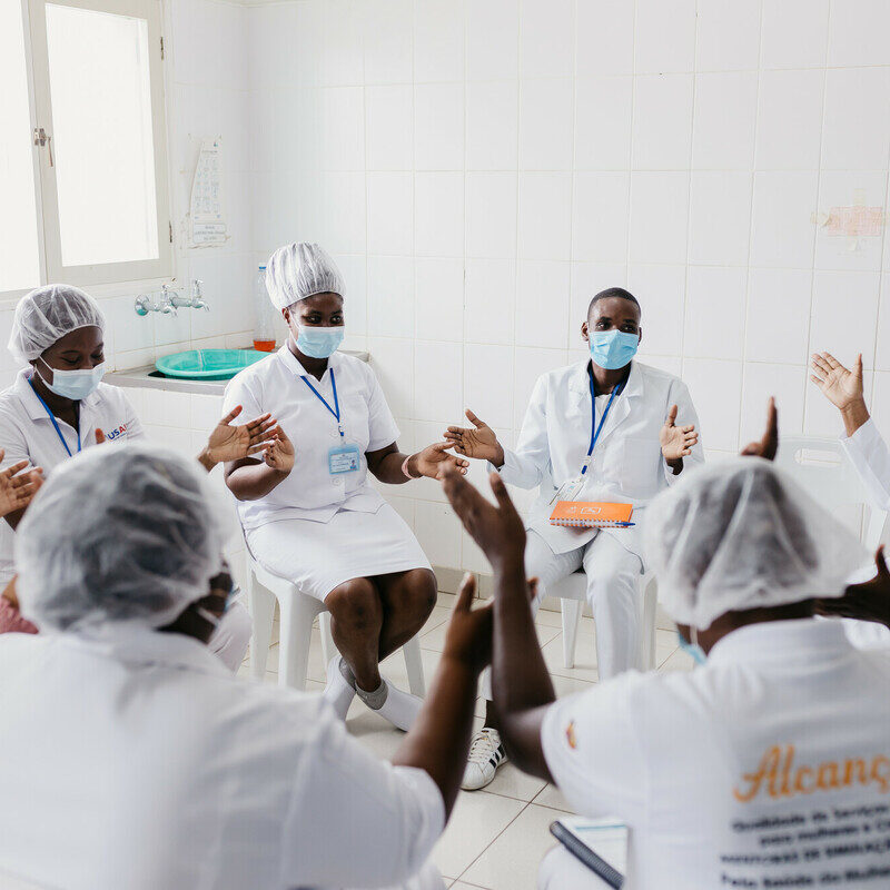 A group of nurses wearing face masks and white dresses with "Alcancar" printed on the back sit in a circle and clap.
