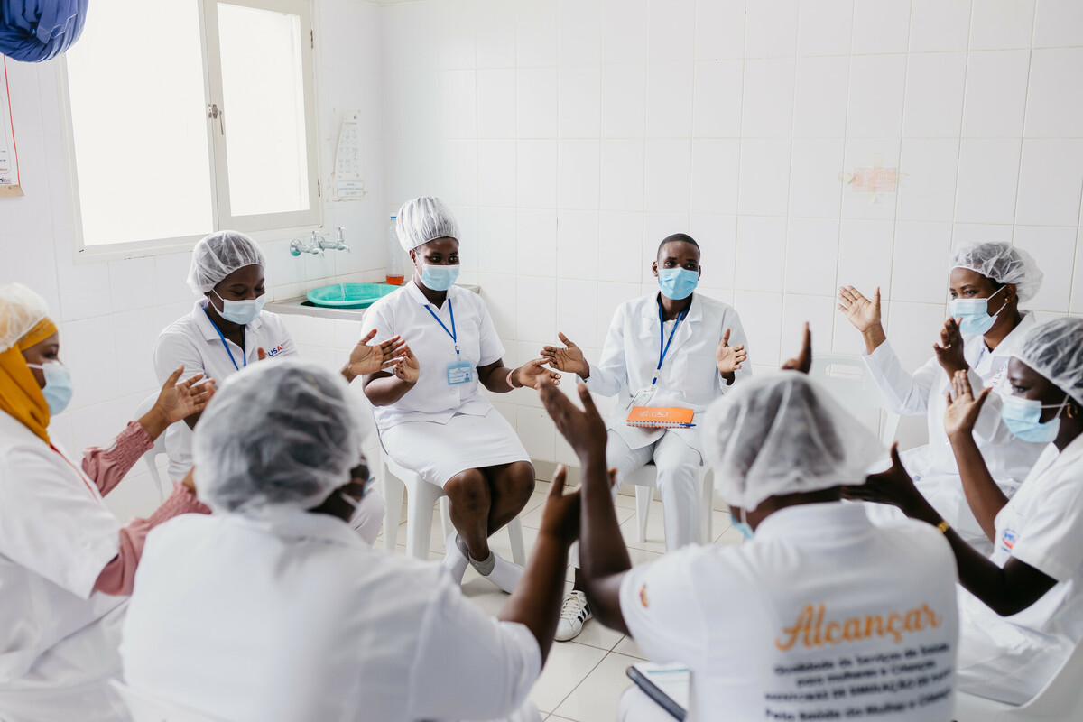 A group of nurses wearing face masks and white dresses with "Alcancar" printed on the back sit in a circle and clap.
