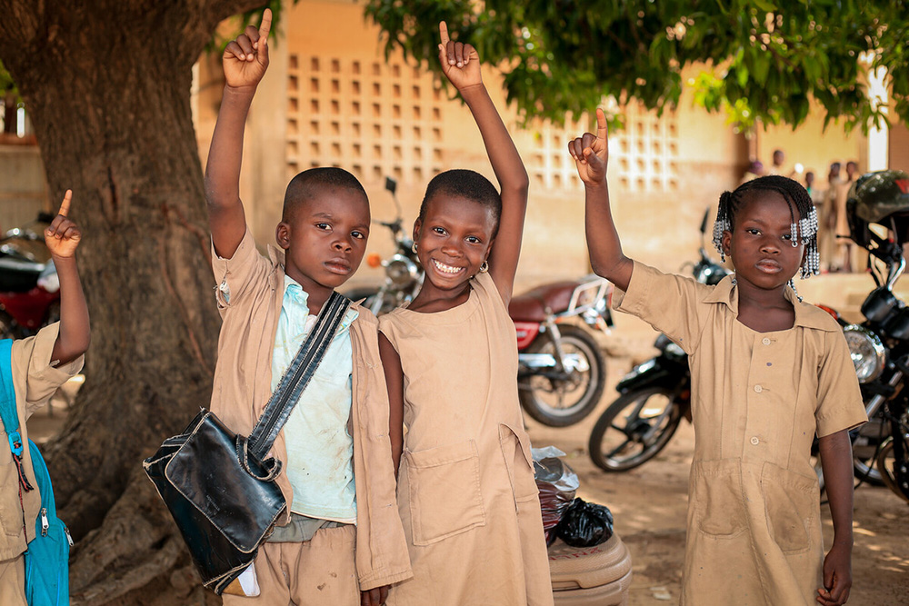 3 young kids wearing school uniforms stand with their hands raised outside their school building.