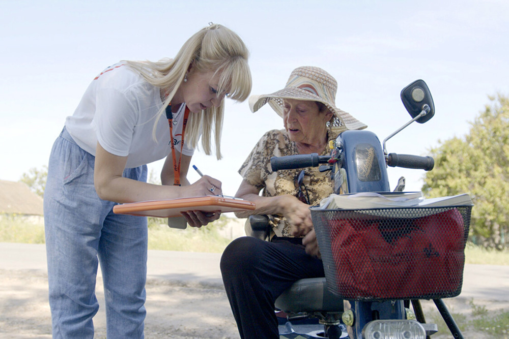 A woman wearing an FHI 360 lanyard and t-shirt writes information on a clipboard while speaking to a older woman in a motorized wheelchair.