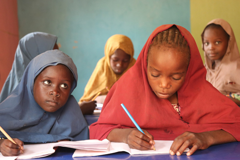 Young girls wearing colorful head coverings write in their notebooks in a classroom.