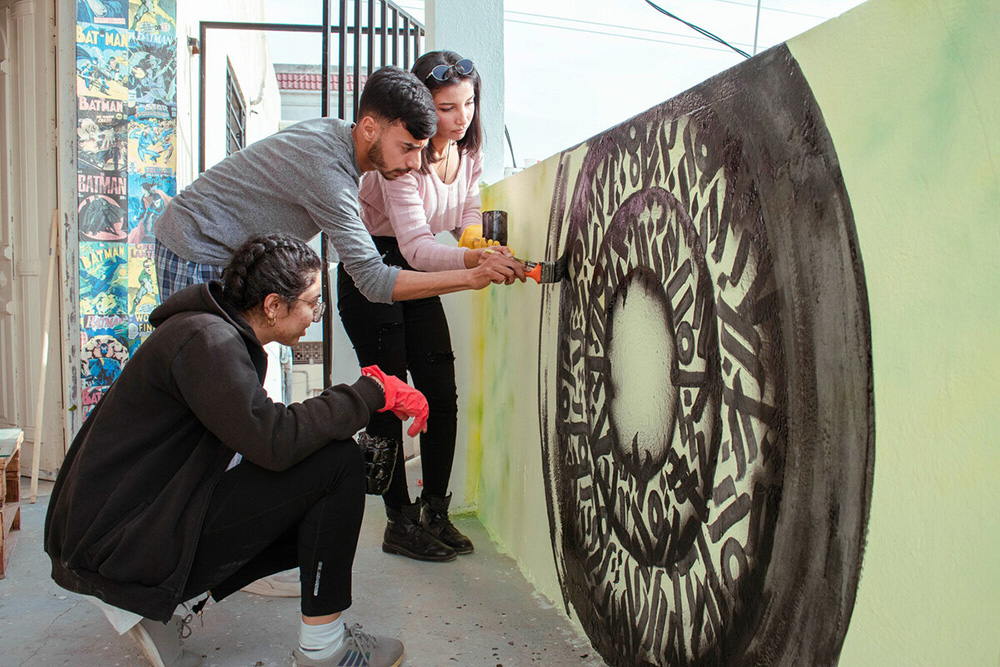 Three young people paint a mural on a concrete wall.