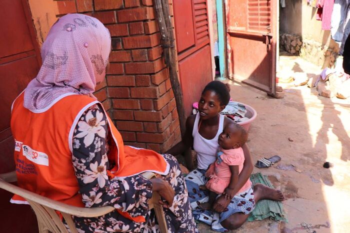 A woman sits on the ground with her baby on her lap and speaks to a community health worker sitting in a chair.