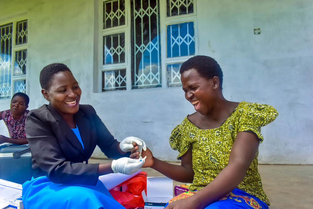Two women laugh while one takes a small blood sample from the other for testing.