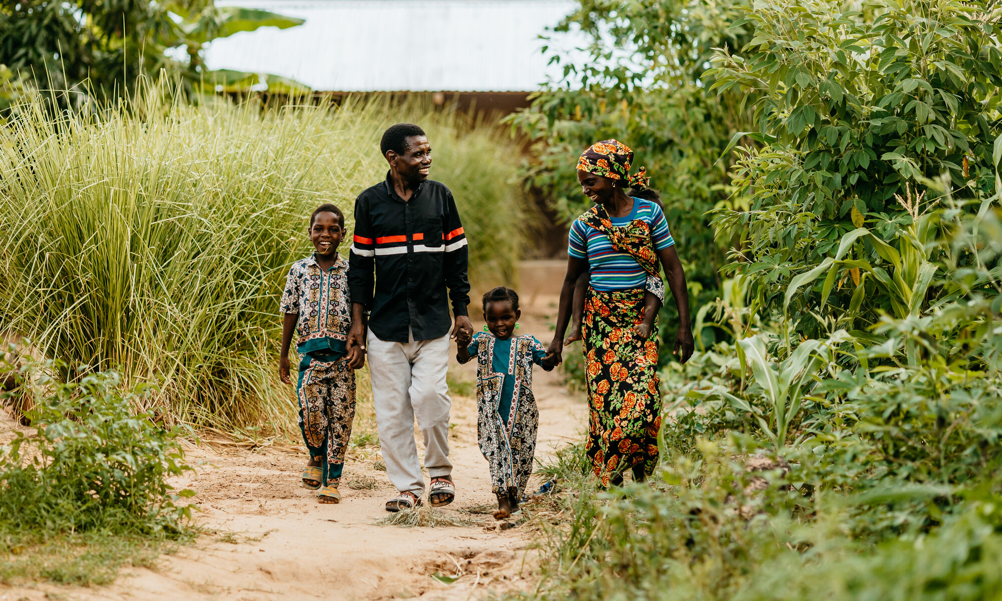 A smiling family holding hands walk down a dirt path through foliage.
