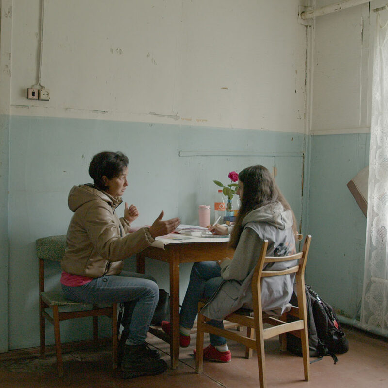 Two women sit at a small table inside a room. White lace curtains cover the window, and the walls are painted white and light blue.