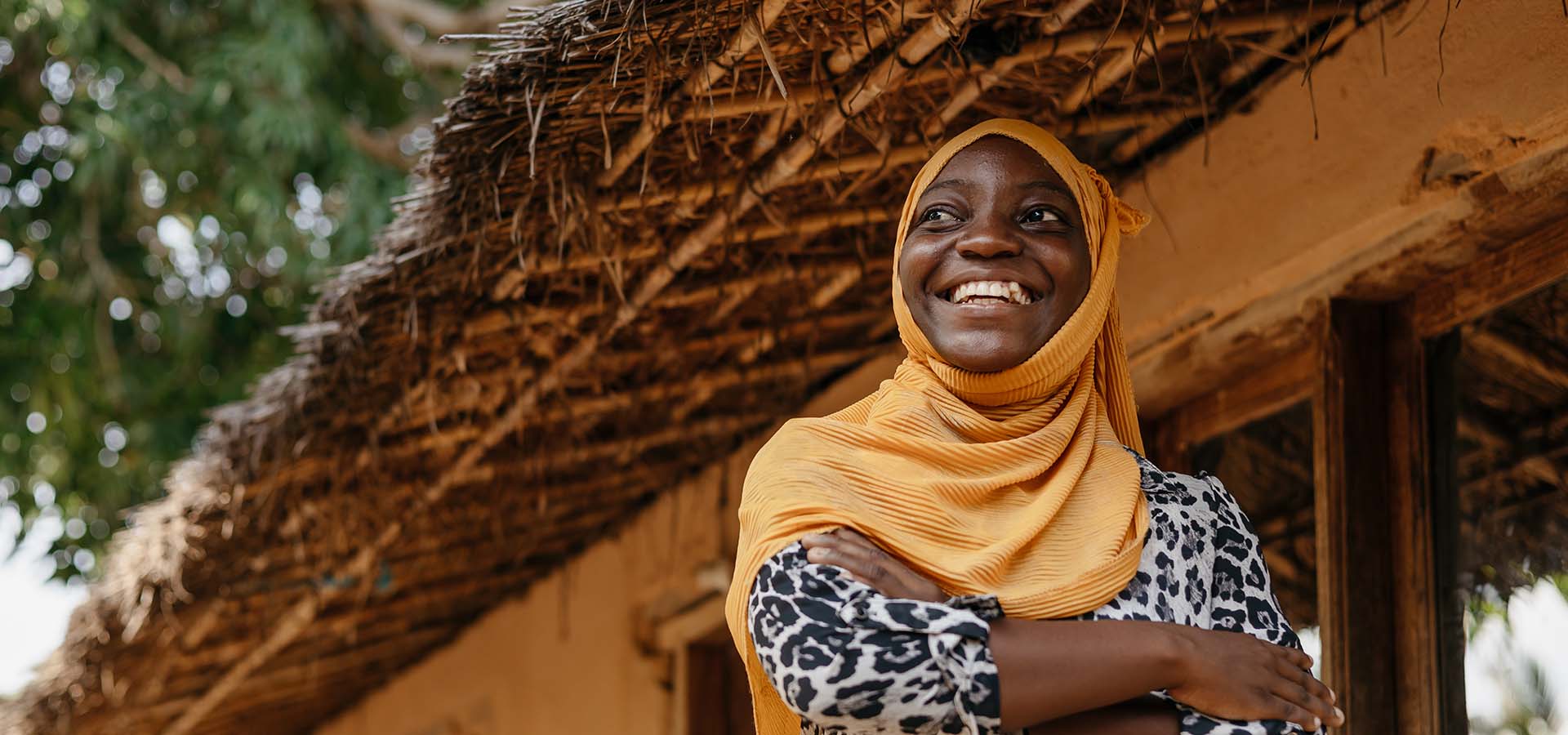 A young woman in a yellow headscarf laughs while standing under the awning of her home.
