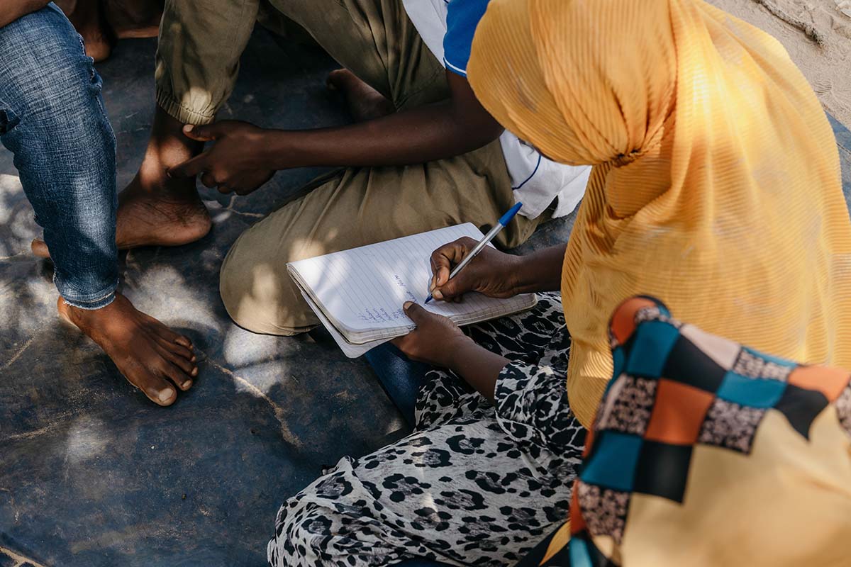 The young woman is seated on the ground in a group of people. She's writing in a notebook.