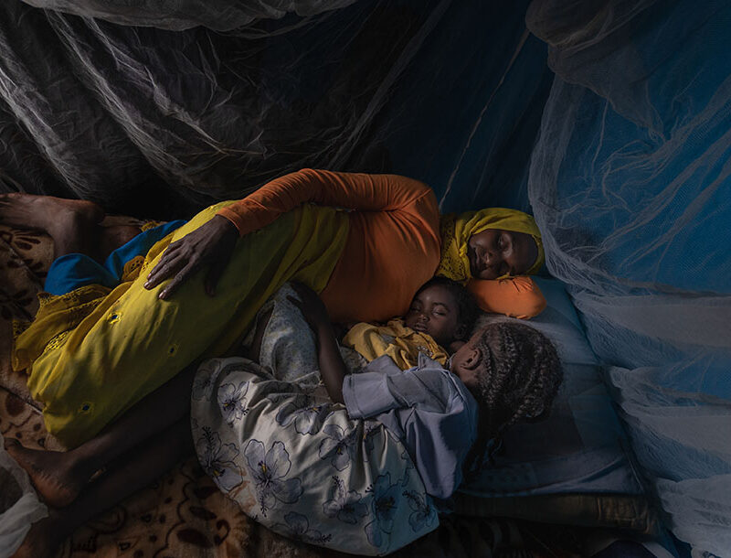 A woman and her two children lay side by side sleeping under a white mosquito net.
