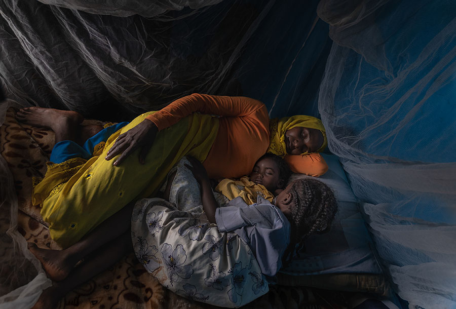 A woman and her two children lay side by side sleeping under a white mosquito net.