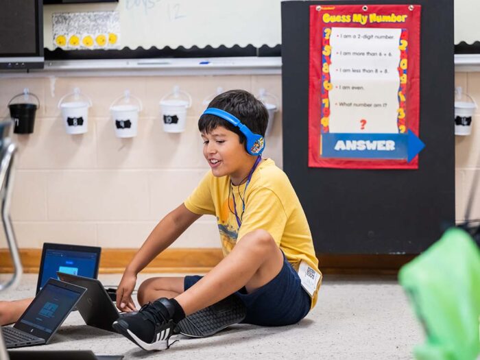 A boy in a yellow t-shirt and blue headphones smiles while sitting on the floor and using a laptop.