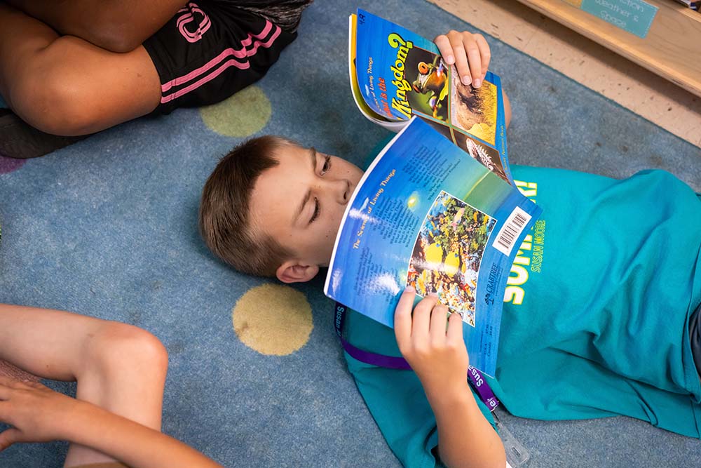 A boy lays on his back on a blue and yellow polka dot carpet, engrossed in a book about animals.