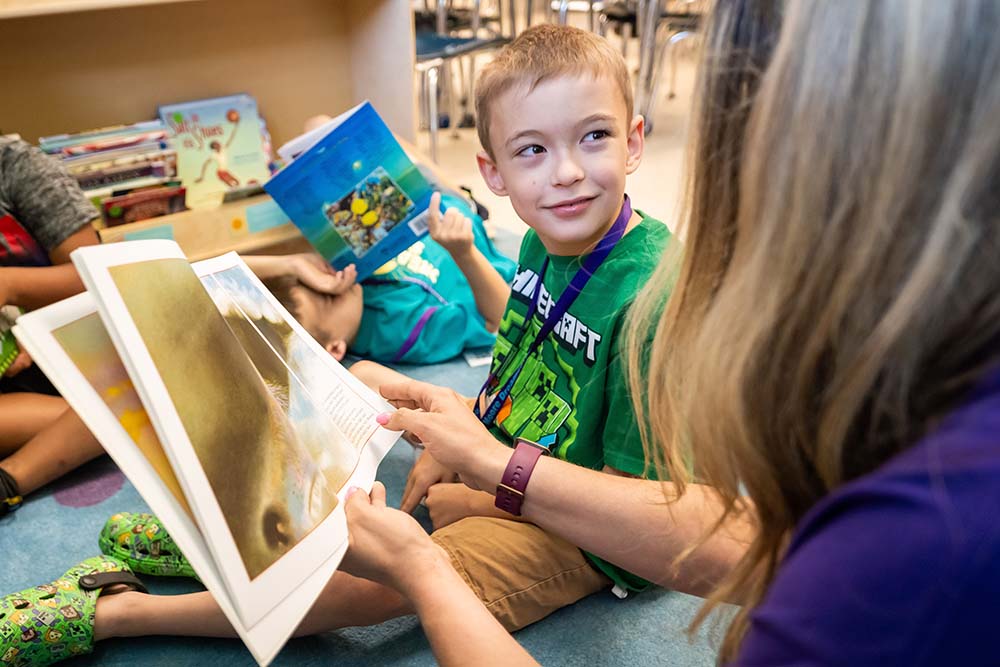 A student in a green t-shirt sits on the floor and smiles at his teacher as they both look through a book.