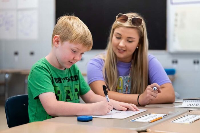 A boy in a green shirt writes while seated next a teacher, who is helping him.