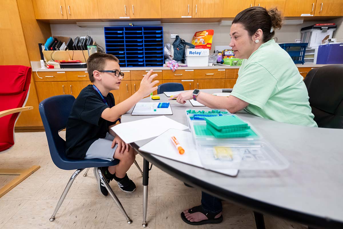 A young boy in a black t-shirt and glasses sits across a table from a teacher as they discuss a lesson.