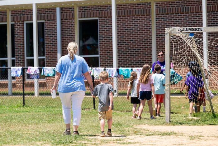 A group of kids walk through a soccer field. In the foreground, an instructor holds the hand of a young boy in a gray t-shirt.