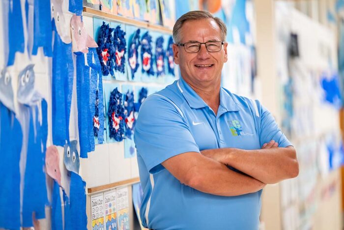 A man wearing a light blue polo shirt smiles and crosses his arms in the hallway of an elementary school.