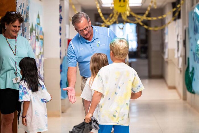 A man sticks his hand out to receive high 5's from a line of kids as they enter a classroom.