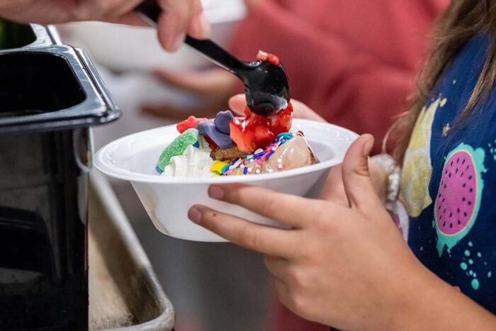 A student holds a bowl with ice cream, while an adult scoops strawberries on top.